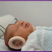 A newborn baby is being tested with a white adhesive pad placed around the head and neck, surrounding the ear, to check their response to sound.